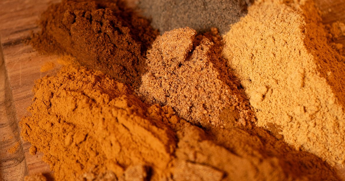 Organic chai powder in a small bowl beside whole cardamom pods, cinnamon sticks, and black peppercorns on a rustic surface